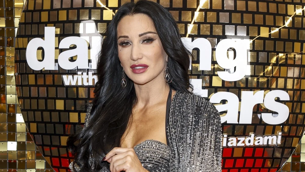 Kobieta o długich ciemnych włosach, ubrana w srebrzystą kreację, stoi na tle złotej, lustrzanej ściany ozdobionej logotypem programu Dancing with the Stars. Sylwetka podkreślona eleganckim makijażem i wyrazistymi ustami.