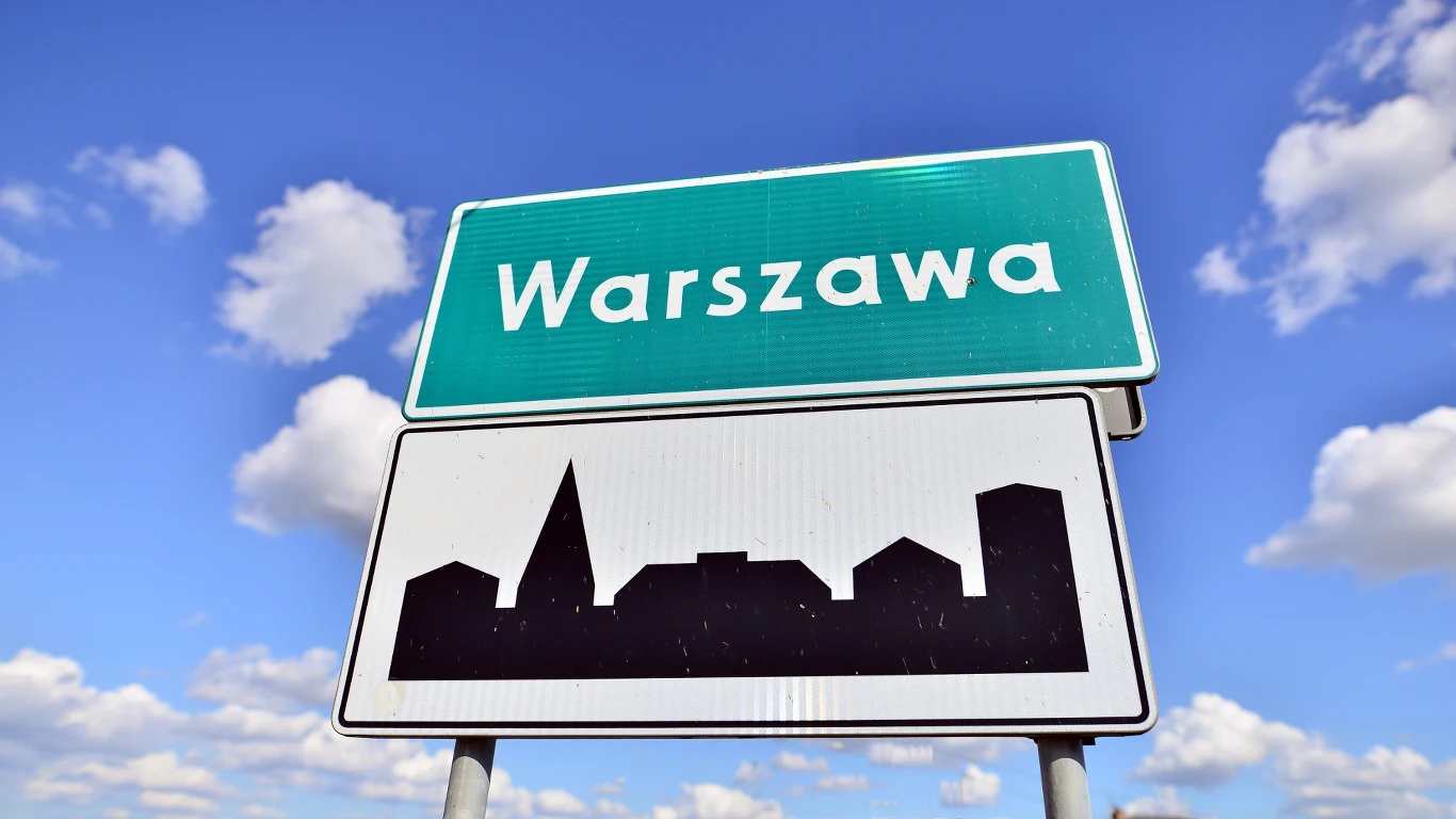 Czy w Warszawie usłyszymy regionalizmy? Zielona tablica z białym napisem Warszawa oraz biała tablica z czarnym konturem miejskiej panoramy na tle niebieskiego nieba z białymi chmurami.