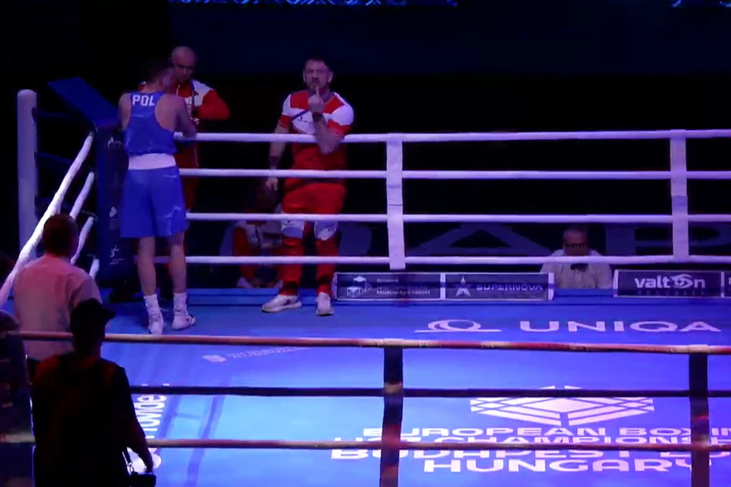 Dwóch bokserów na ringu podczas przerwy w walce, jeden ubrany w niebieski strój z napisem POL, drugi w czerwonym stroju. Trenerzy lub sędziowie obecni w rogach ringu, ring otoczony przez białe liny, dobrze oświetlone miejsce zawodów sportowych.