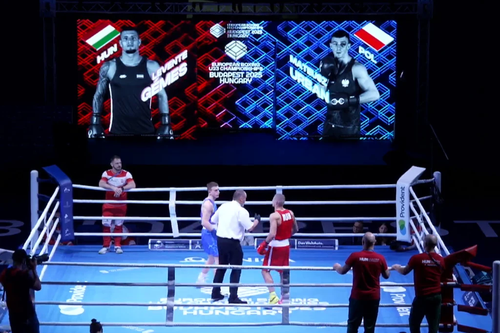 Dwóch bokserów w ringu przygotowuje się do walki, sędzia stoi pomiędzy nimi, a w tle znajduje się duży ekran z wizualizacjami zawodników z Węgier i Polski oraz informacjami o turnieju bokserskim.