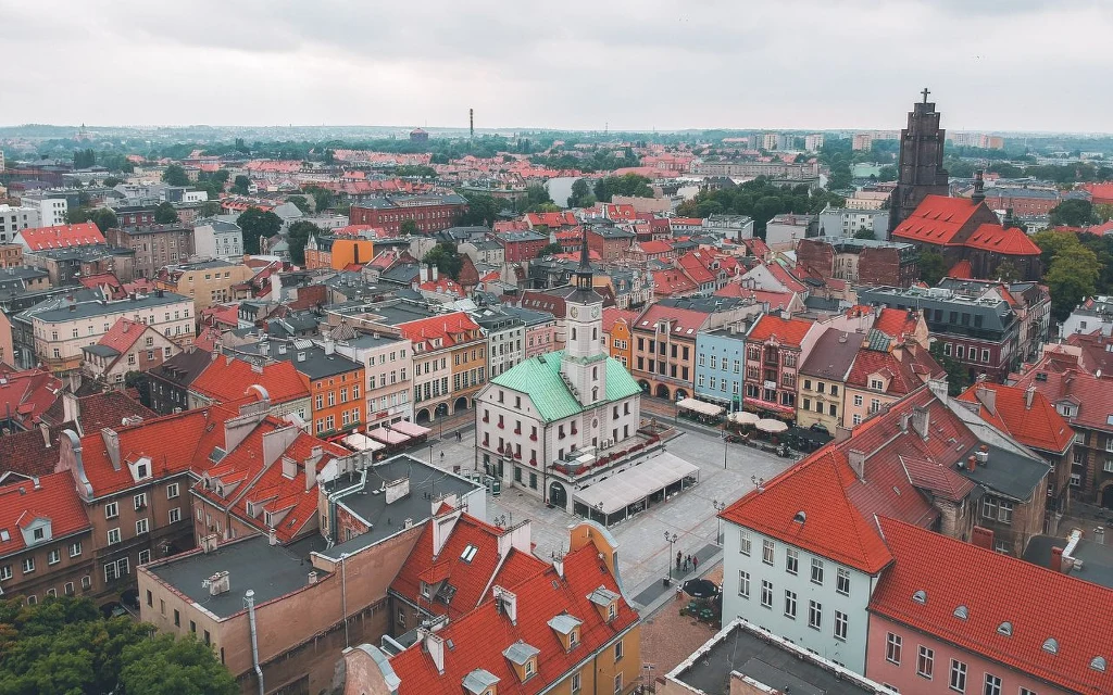 Rynek miejski otoczony kamienicami z czerwonymi dachami, centralnie usytuowany budynek ratusza z zielonym dachem, brukowane ulice i rozpościerające się na horyzoncie miejskie zabudowania.