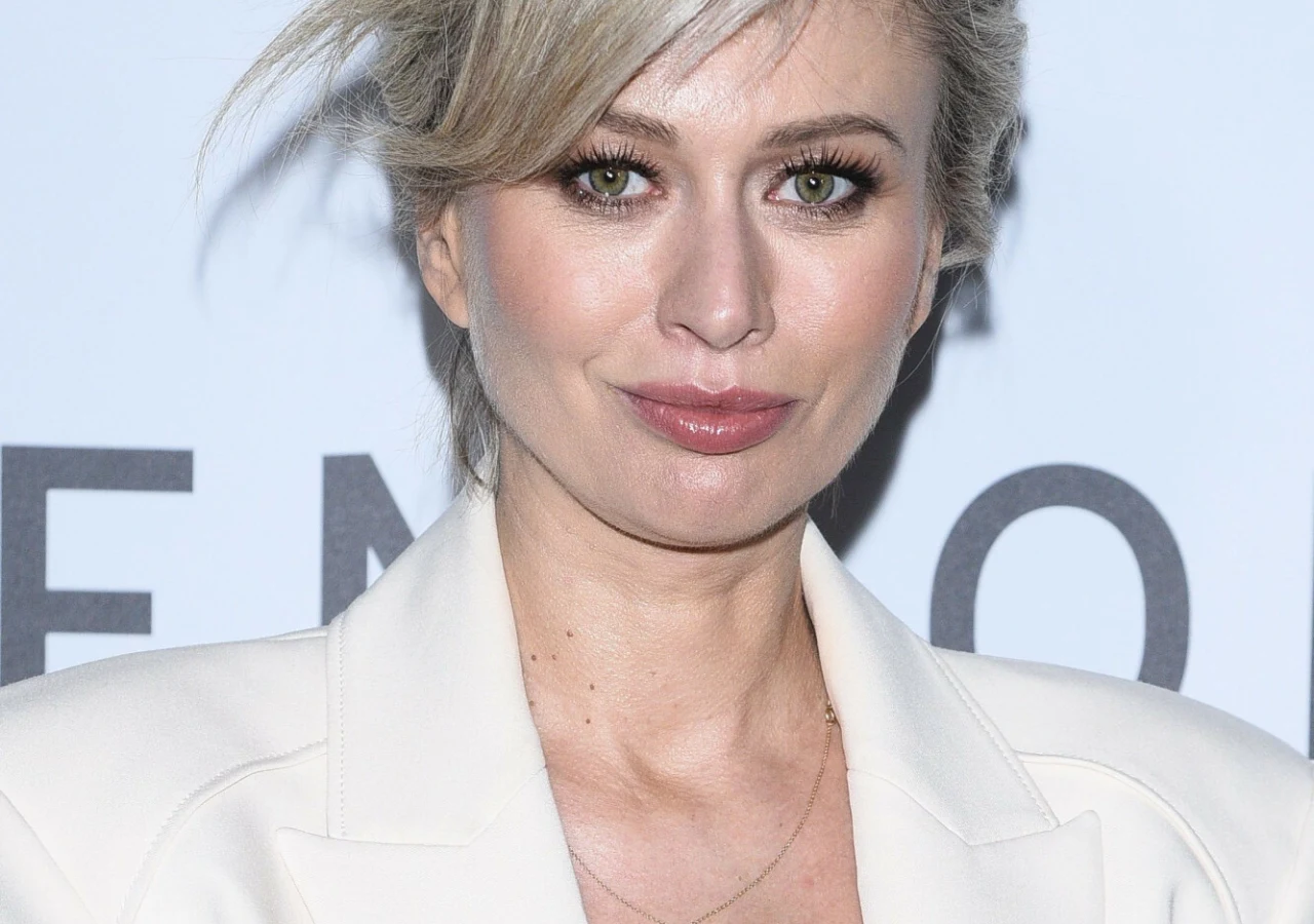 Katarzyna Zdanowicz Kobieta o blond włosach i zielonych oczach ubrana w elegancką białą marynarkę na tle jasnej ścianki z elementami tekstu.