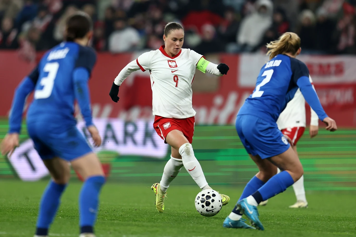 Piłkarka w białym stroju z numerem 9 drybluje piłką podczas meczu, otoczona przez dwie zawodniczki drużyny przeciwnej ubrane na niebiesko. W tle widoczni kibice i stadionowe oświetlenie.