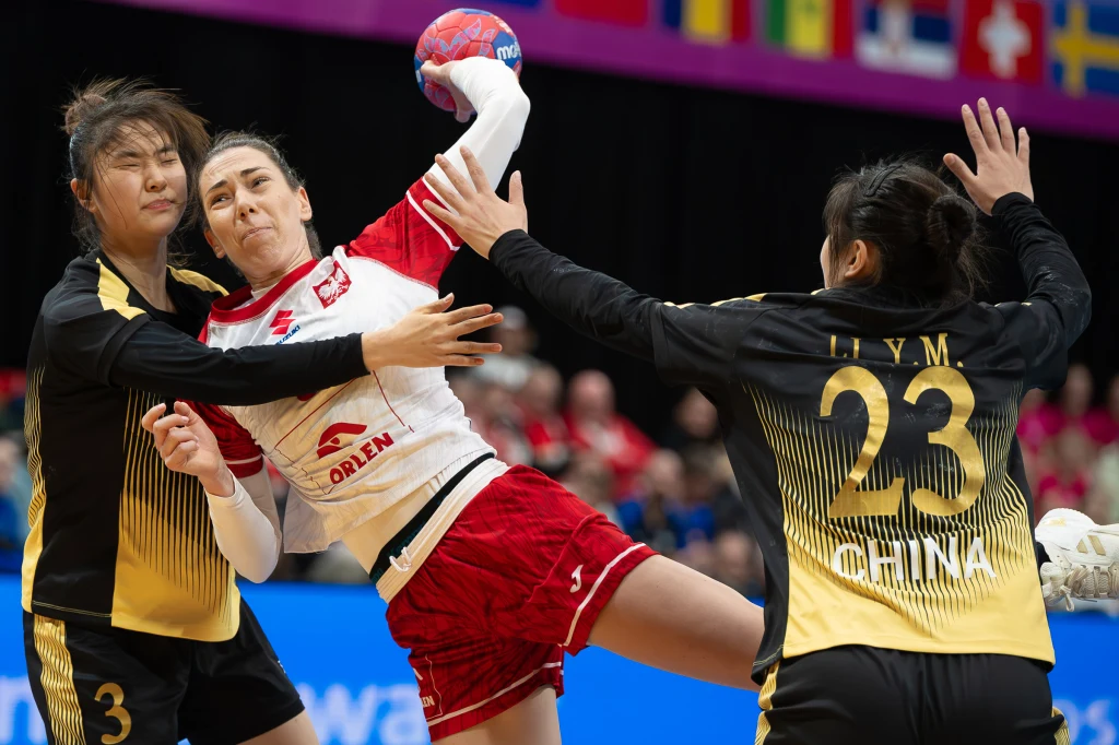 Dynamiczna akcja podczas meczu piłki ręcznej kobiet, jedna z zawodniczek w biało-czerwonym stroju próbuje rzucić piłkę, będąc bronioną przez dwie zawodniczki w żółto-czarnych strojach z drużyny Chin.