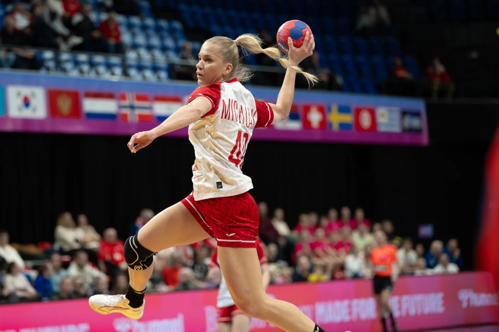 Zawodniczka w biało-czerwonym stroju piłki ręcznej wykonuje dynamiczny rzut w powietrzu na tle hali sportowej z widownią, za nią widoczne są flagi narodowe oraz inni uczestnicy meczu.