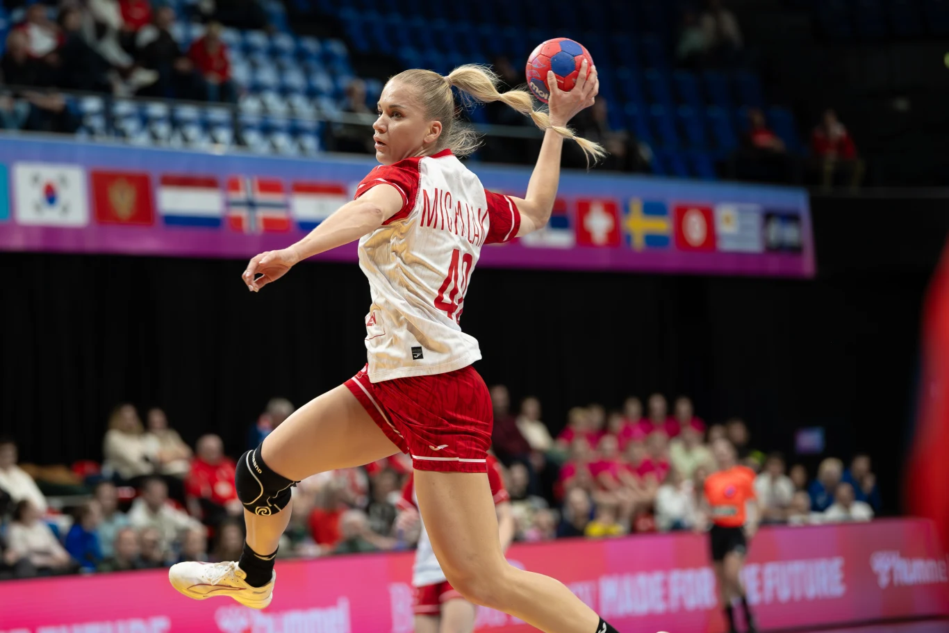 Zawodniczka w biało-czerwonym stroju piłki ręcznej wykonuje dynamiczny rzut w powietrzu na tle hali sportowej z widownią, za nią widoczne są flagi narodowe oraz inni uczestnicy meczu.