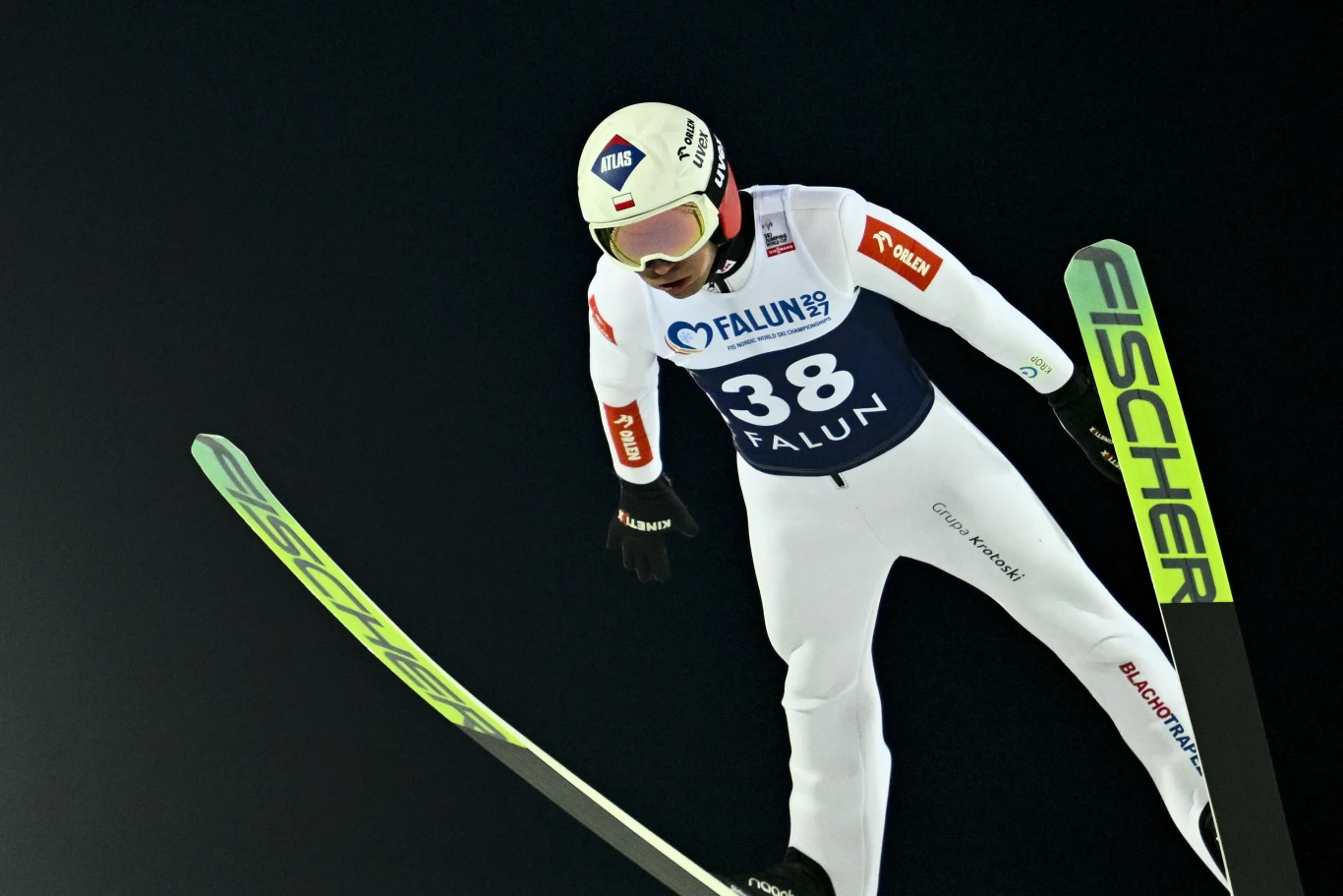 Kamil Stoch Skoczek narciarski w trakcie lotu na nartach w pełnym stroju sportowym, wyposażony w kask i kombinezon, na tle ciemnego nieba, numer startowy 38, widoczna marka nart FISCHER oraz logo Falun 2021.