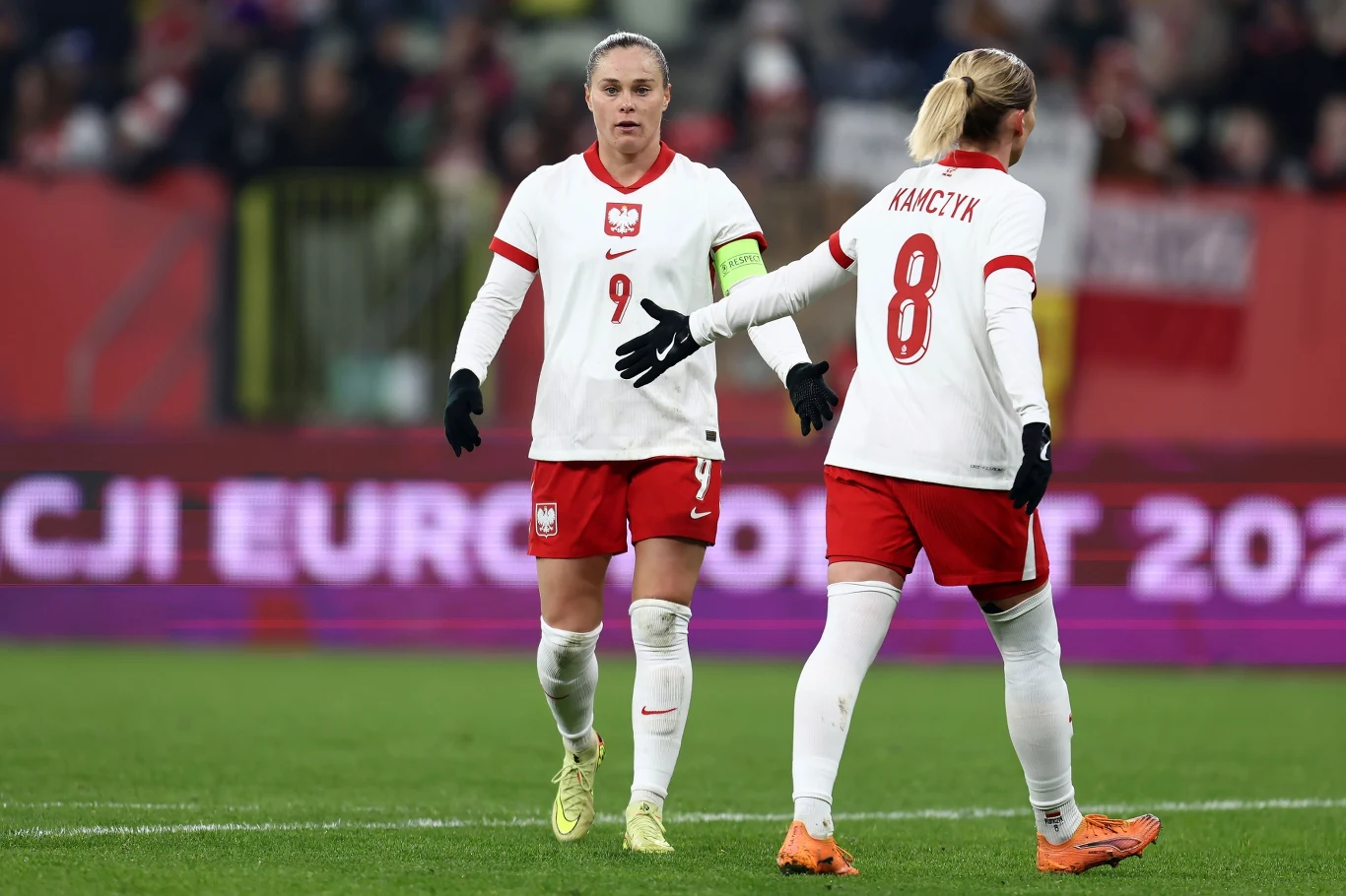 Dwie polskie piłkarki w białych koszulkach i czerwonych spodenkach wymieniają gesty na boisku, widoczne numery 9 oraz 8 na strojach, w tle rozmyta publiczność i fragmenty stadionu.