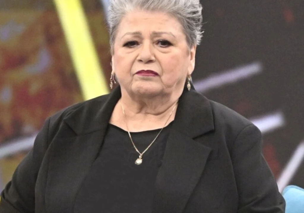 Kobieta o siwych włosach ułożonych do góry, ubrana w czarną marynarkę oraz naszyjnik z zawieszką, siedzi na tle rozmytego, wielokolorowego tła z żółtym i czarnym elementem.