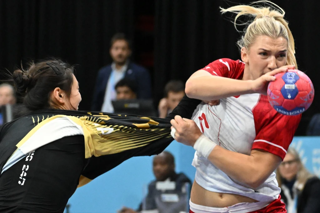 Dwóch zawodniczek walczy o piłkę ręczną, jedna z nich ciągnie przeciwniczkę za koszulkę podczas dynamicznej akcji na tle rozmazanej publiczności i czarnego tła.