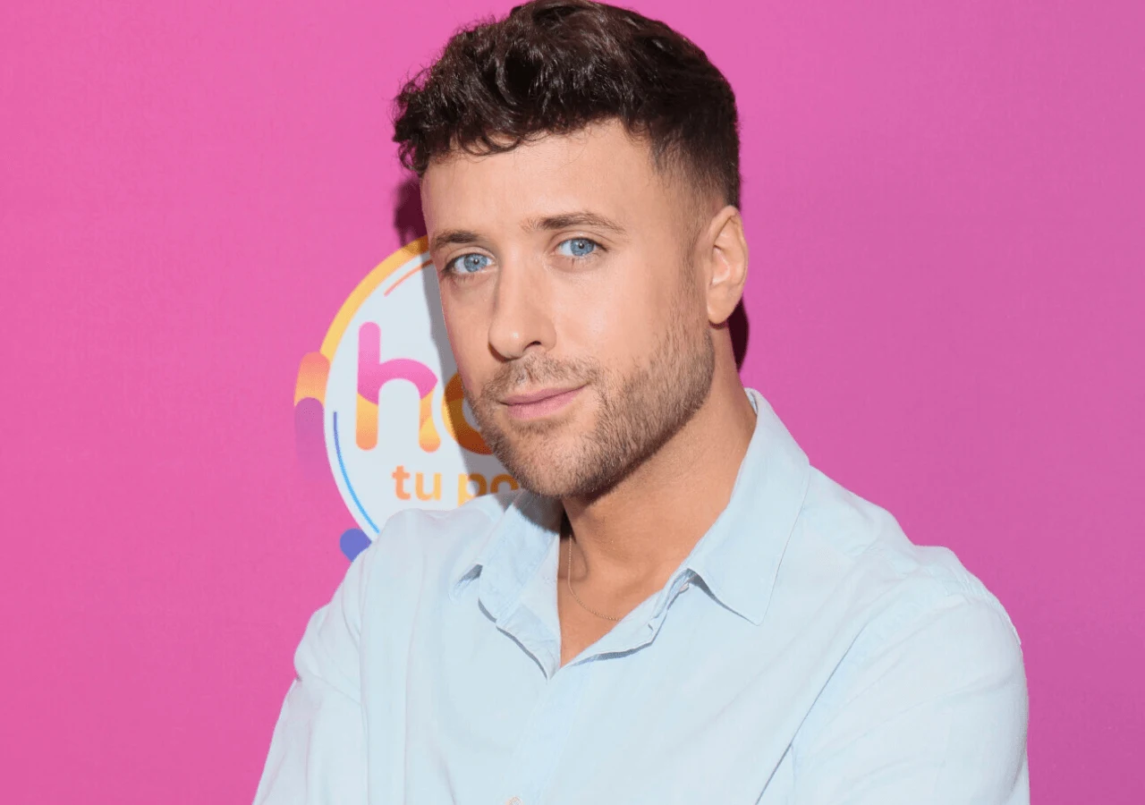 Aleksander Sikora Mężczyzna z krótkimi, ciemnymi włosami i jasnoniebieskimi oczami ubrany w błękitną koszulę pozuje na tle różowej ściany z kolorowym logo w tle.