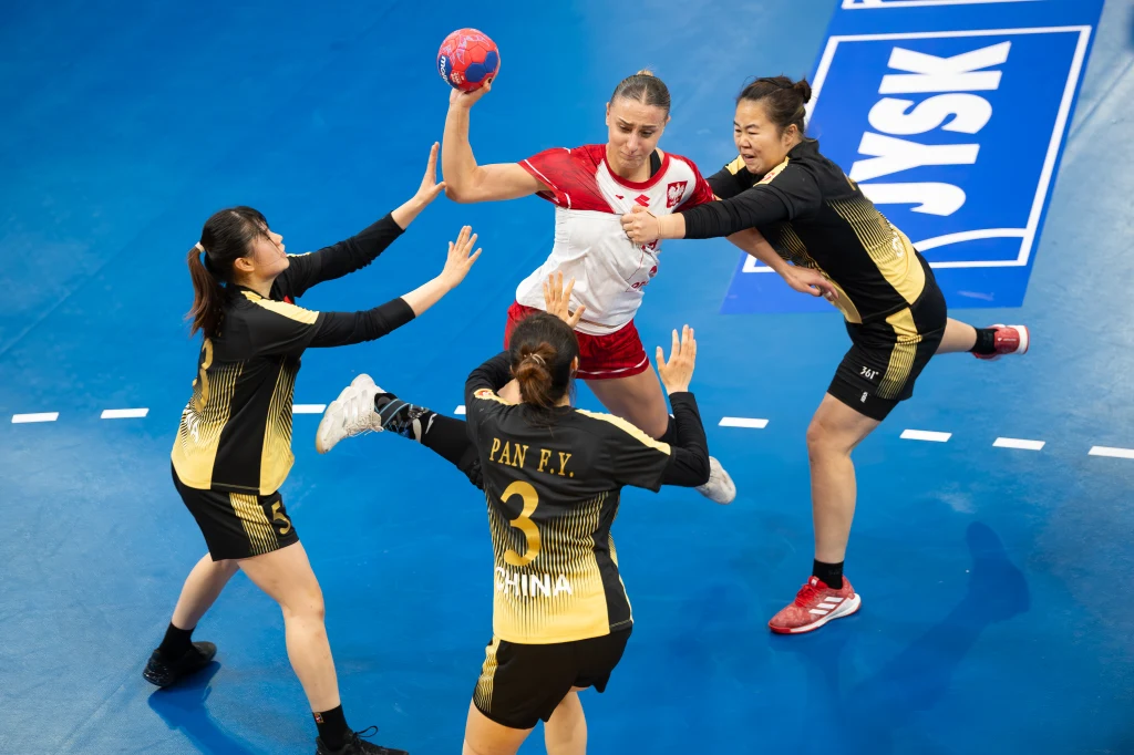 Zawodniczka w biało-czerwonym stroju z piłką w ręce próbuje przedrzeć się przez obronę trzech zawodniczek w czarno-żółtych strojach z napisem 'CHINA' na niebieskim boisku piłki ręcznej.