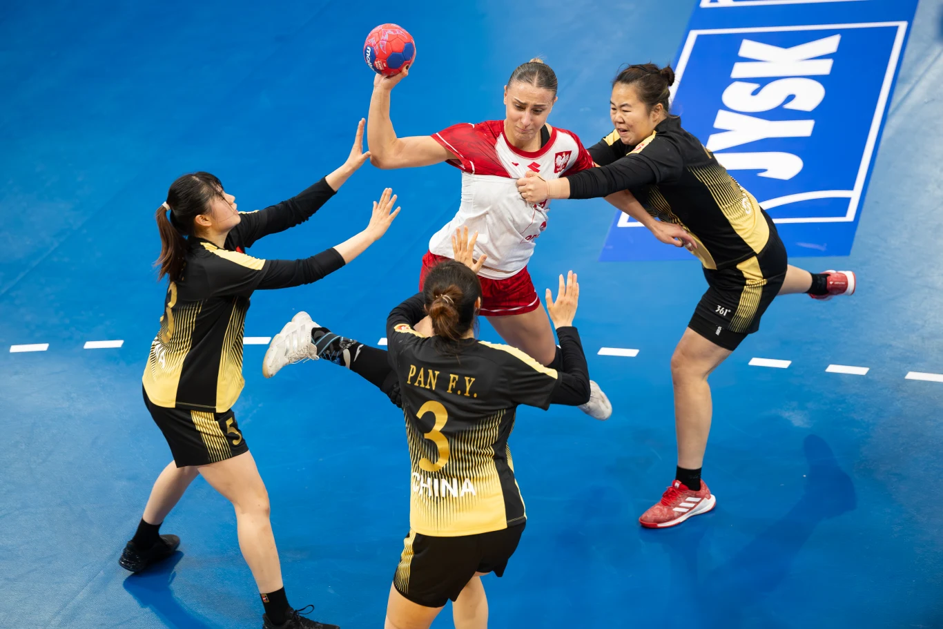 Zawodniczka w biało-czerwonym stroju z piłką w ręce próbuje przedrzeć się przez obronę trzech zawodniczek w czarno-żółtych strojach z napisem 'CHINA' na niebieskim boisku piłki ręcznej.
