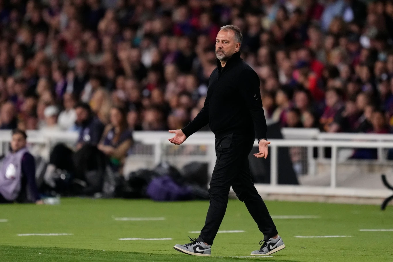 Mężczyzna ubrany na czarno spaceruje po boisku sportowym podczas meczu piłki nożnej, w tle widoczna jest rozmyta publiczność na trybunach.