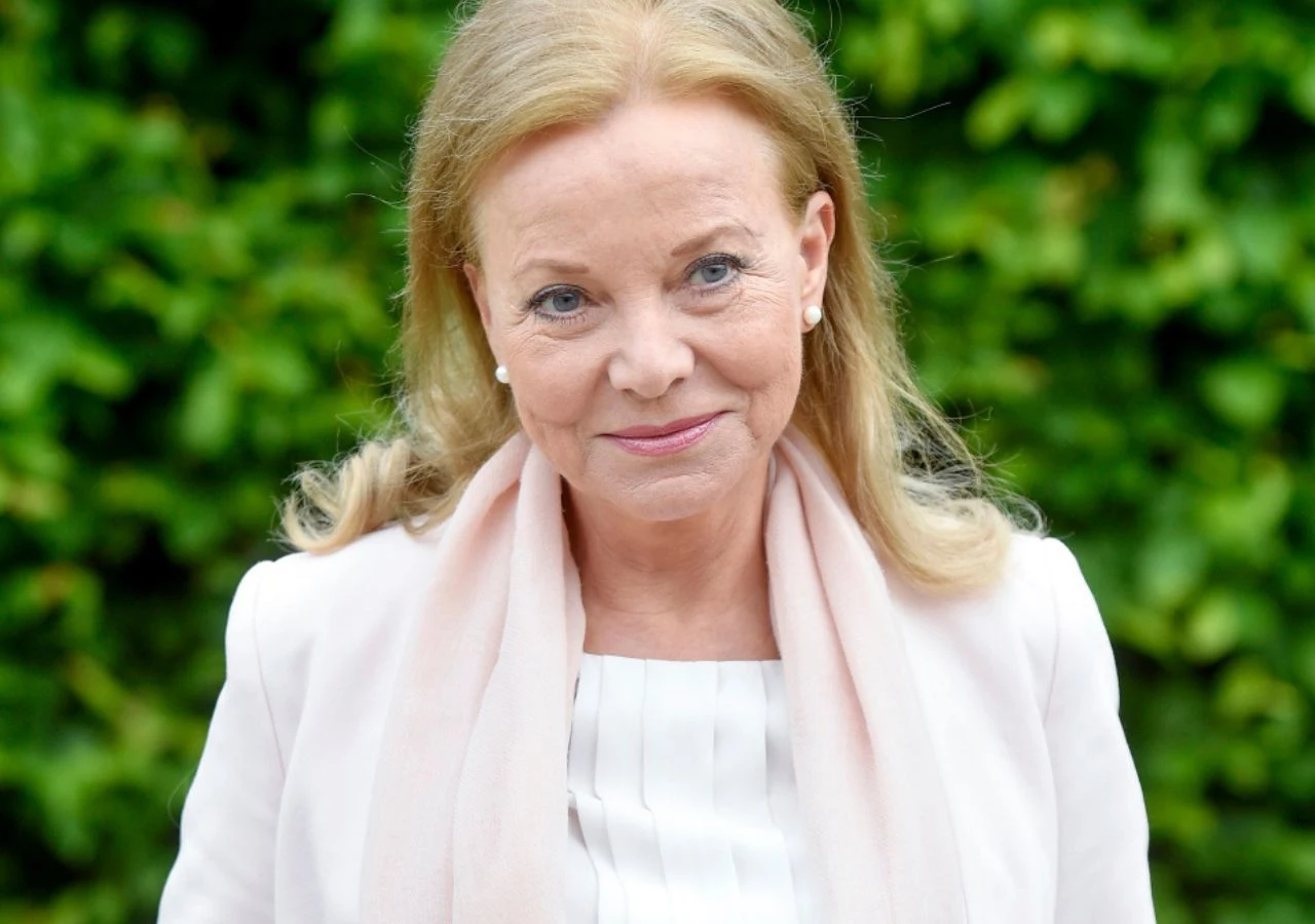 Laura Łącz Starsza kobieta z blond włosami uśmiecha się delikatnie, ubrana w jasny żakiet i szalik, na tle zielonych, rozmytych liści.