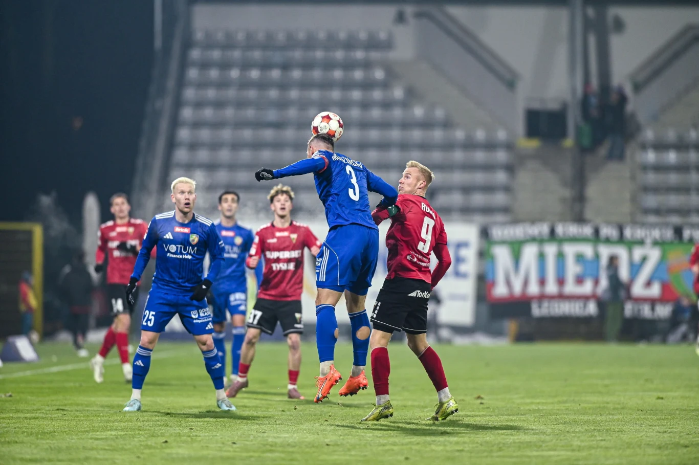 Zawodnik drużyny w niebieskich strojach wyskakuje do piłki głową podczas meczu piłkarskiego. Otaczają go gracze obu drużyn w niebieskich i bordowych koszulkach, a w tle widoczne są puste trybuny oraz transparent z nazwą klubu.