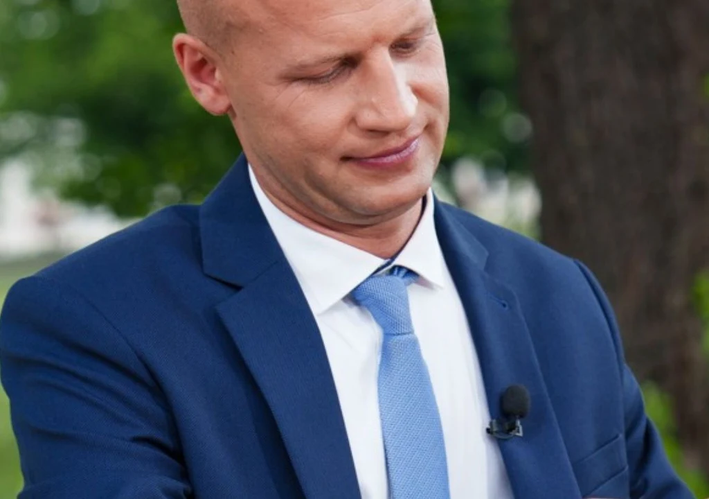 Mężczyzna w garniturze i niebieskim krawacie z mikrofonem przypiętym do klapy marynarki w plenerze, z wyrazem skupienia na twarzy.