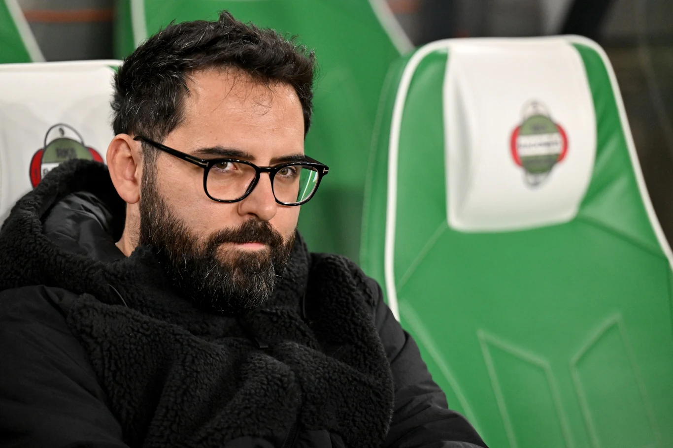 Mężczyzna z brodą i okularami siedzi na zielonym fotelu stadionowym, ubrany w czarne ubranie zimowe, patrzy poważnie w stronę aparatu, tło stanowią puste siedzenia.