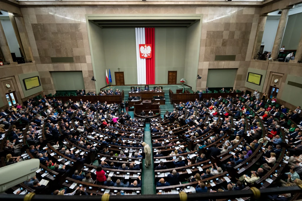 Sondaż. Czy Polacy chcą nowej partii politycznej? (zdj. ilustracyjne) Sala plenarna polskiego parlamentu wypełniona przez licznie zgromadzonych posłów i posłanki, na ścianie centralnie zawieszona flaga Polski z godłem, po bokach flagi Polski i Unii Europejskiej, półokrągły układ miejsc dla deputowanych.