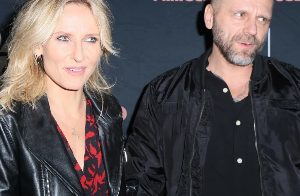 Patrycja Markowska z mężem Kobieta o blond włosach w czarnej kurtce skórzanej oraz mężczyzna z zarostem ubrany w ciemną kurtkę stoją razem na tle czarnej ściany z napisem.