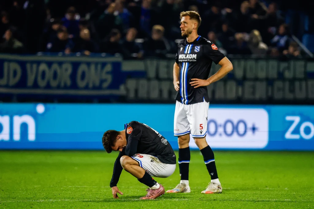 Dwóch piłkarzy w czarnych strojach na boisku, jeden stoi z rękami na biodrach i patrzy przed siebie, drugi kuca ze spuszczoną głową, wyrażając rozczarowanie po meczu, w tle rozmazana publiczność i reklamy.