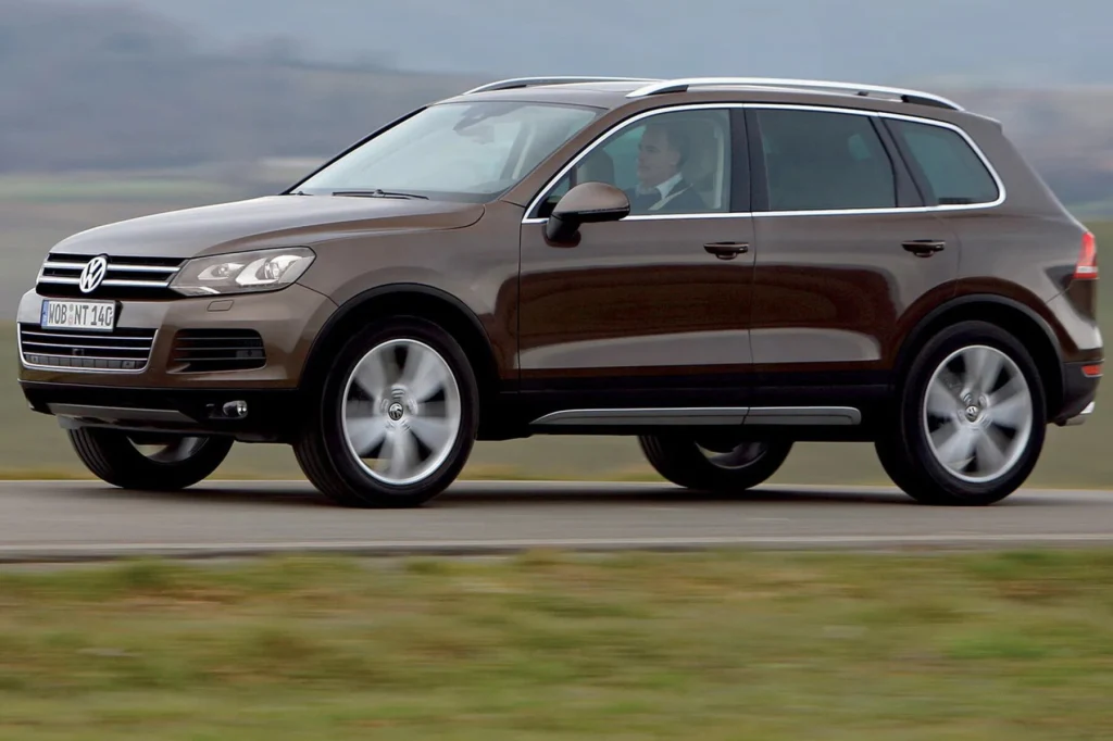 VW Touareg to jedno z najmniej awaryjnych 10-letnich aut według raportu TÜV Brązowy SUV marki Volkswagen porusza się po asfaltowej drodze, w tle rozległy krajobraz o łagodnych barwach, widoczny kierowca w kabinie pojazdu.