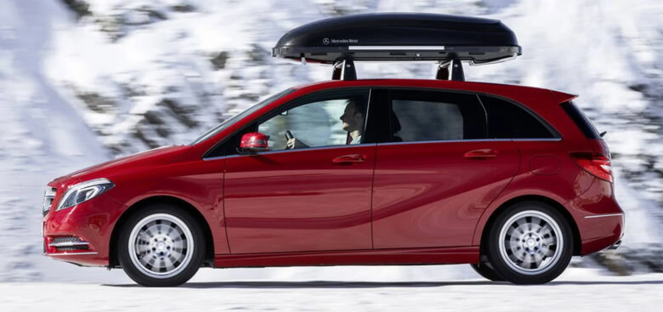 Czerwony samochód osobowy Mercedes z boxem dachowym jedzie po zaśnieżonej drodze na tle zimowego krajobrazu.