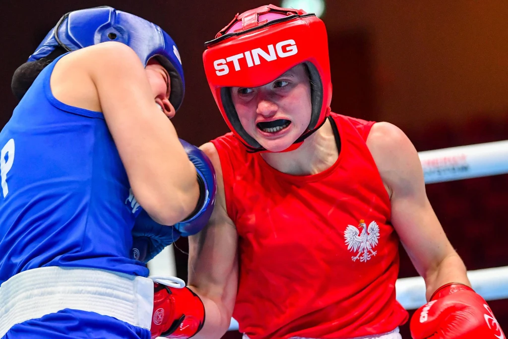 Natalia Kuczewska Dwie zawodniczki boksu rywalizujące na ringu, ubrana w czerwony strój zawodniczka z orłem na piersi wymierza cios przeciwniczce w niebieskim stroju ochronnym, obie w kaskach i ochraniaczach.