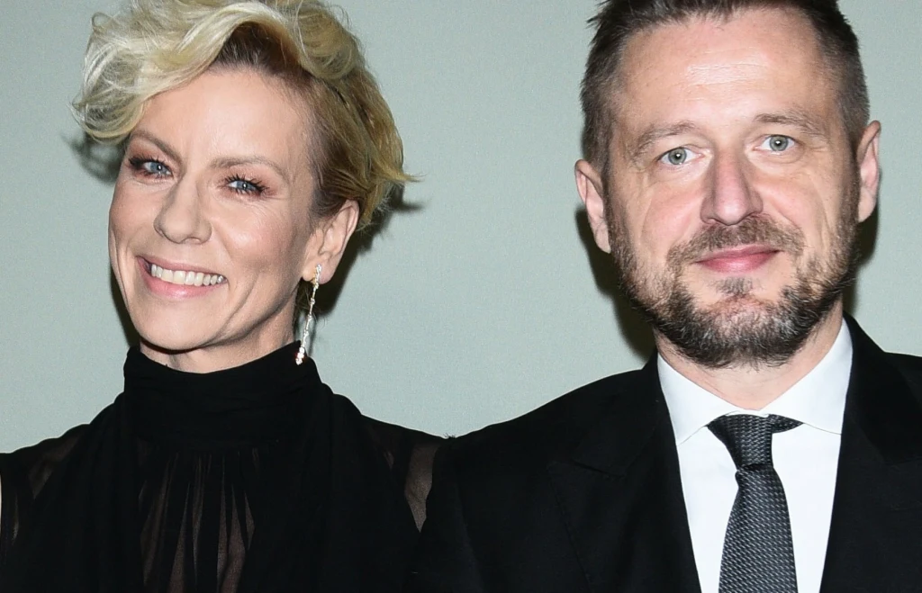 Anita Werner i Michał Kołodziejczyk Dwie elegancko ubrane osoby stojące obok siebie, kobieta z krótkimi blond włosami w czarnej kreacji oraz mężczyzna z brodą w garniturze i krawacie, oboje uśmiechnięci na neutralnym tle.