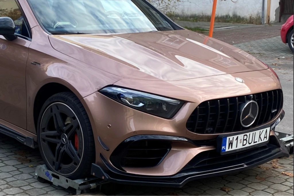 Mercedes AMG wystawiony na licytację przez Urząd Skarbowy w Radomiu. Złoty sportowy samochód marki Mercedes AMG z charakterystyczną tablicą rejestracyjną W1 BULKA, ustawiony na blokadach antykradzieżowych, zaparkowany na brukowanym chodniku przy ulicy.