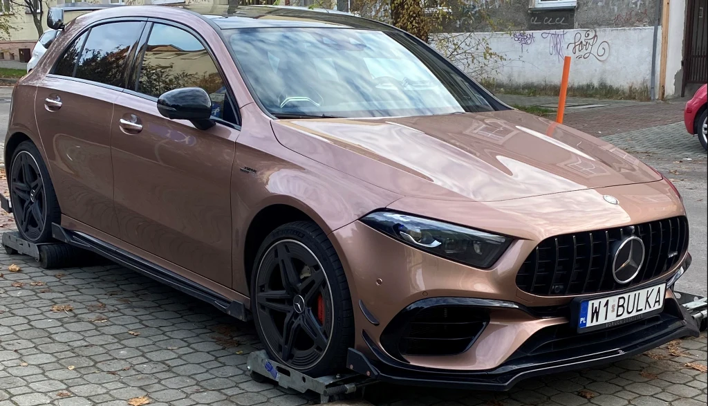 Licytacja odbędzie się 18 grudnia o godzinie 12.00. Brązowy samochód Mercedes klasy A na czarnych felgach, stojący na specjalnych platformach blokujących koła, zaparkowany na bruku w miejskim otoczeniu.