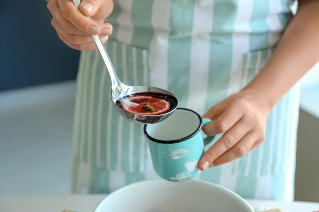 Kompot z suszonych owoców warto pić cały miesiąc. Poprawia pracę jelit i wzmacnia odporność Osoba w fartuchu wlewa zupę lub kompot o ciemnoczerwonym kolorze z chochli do metalowego kubka w jasnym kolorze ozdobionym białymi chmurami.