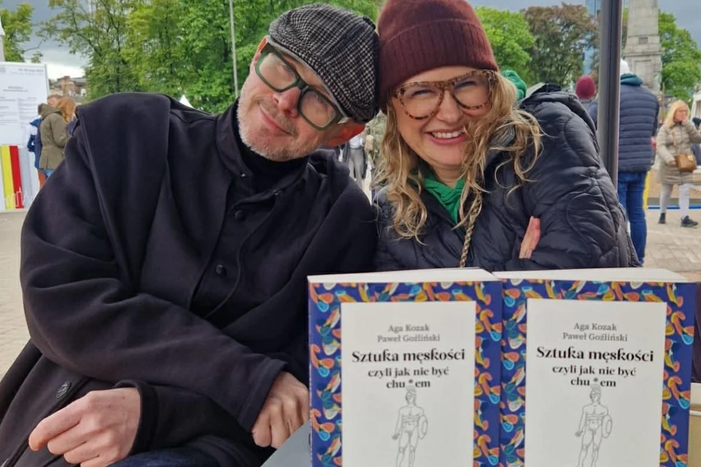 Dwie uśmiechnięte osoby siedzą obok siebie przy stole na świeżym powietrzu, przed nimi leżą dwie książki z tytułem zawierającym wulgaryzm. W tle widać spacerujących ludzi oraz drzewa.