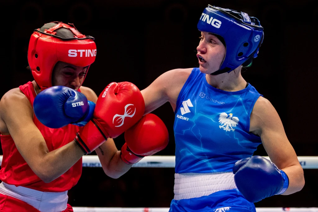 Julia Szeremeta Dwie kobiety w stroju bokserskim walczące na ringu, jedna uderza drugą w głowę, obie mają kaski ochronne i rękawice, widoczna koncentracja i dynamika ruchu.
