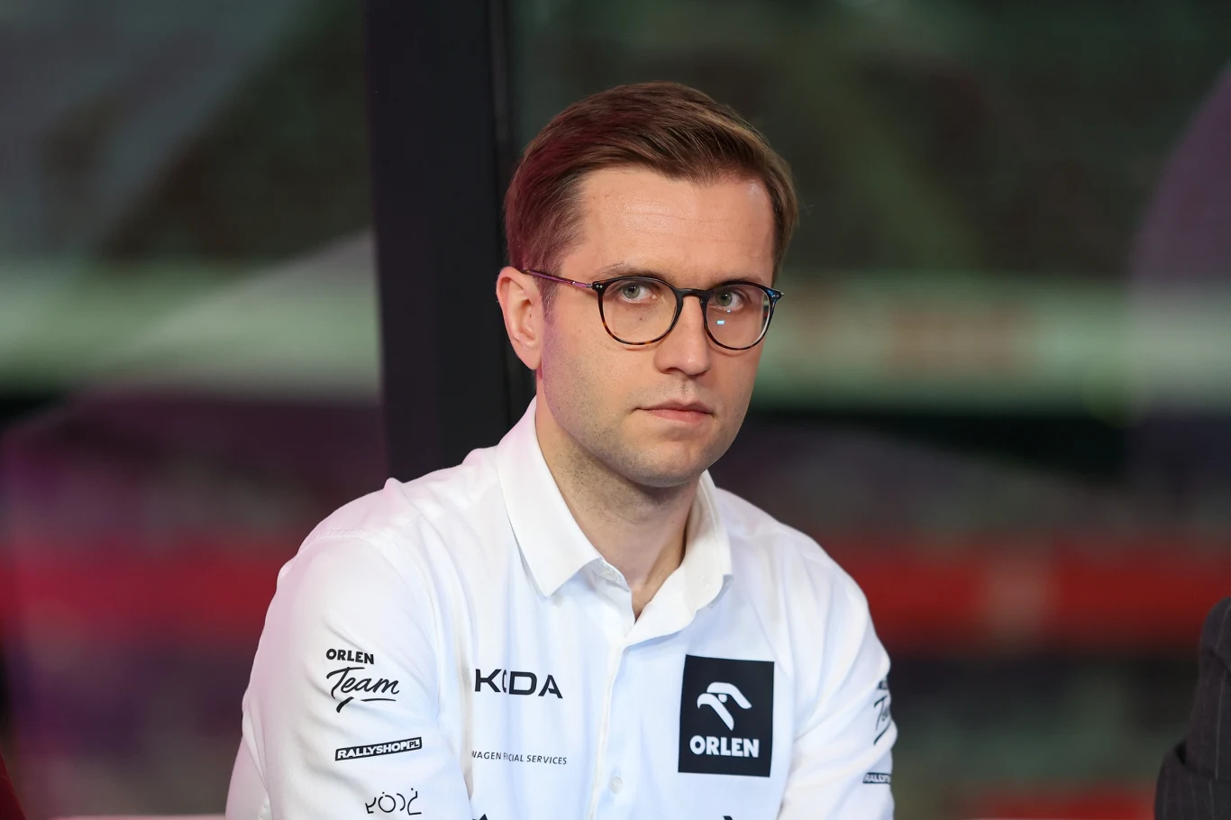 Mikołaj Marczyk Mężczyzna w okularach ubrany w białą koszulę z widocznymi logotypami sponsorów, siedzący we wnętrzu o nowoczesnym charakterze.