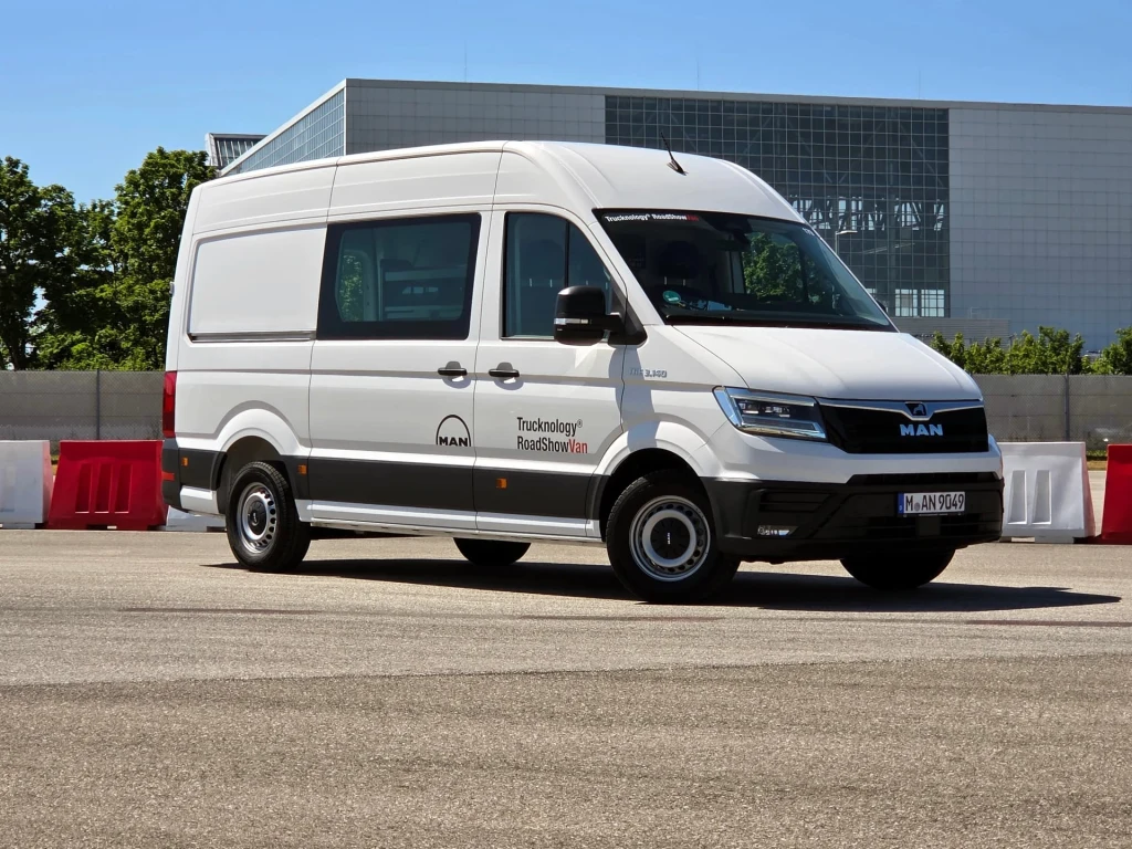 Biały samochód dostawczy marki MAN zaparkowany na asfaltowej powierzchni, w tle nowoczesny budynek z dużymi przeszkleniami oraz betonowy mur i barierki ochronne.