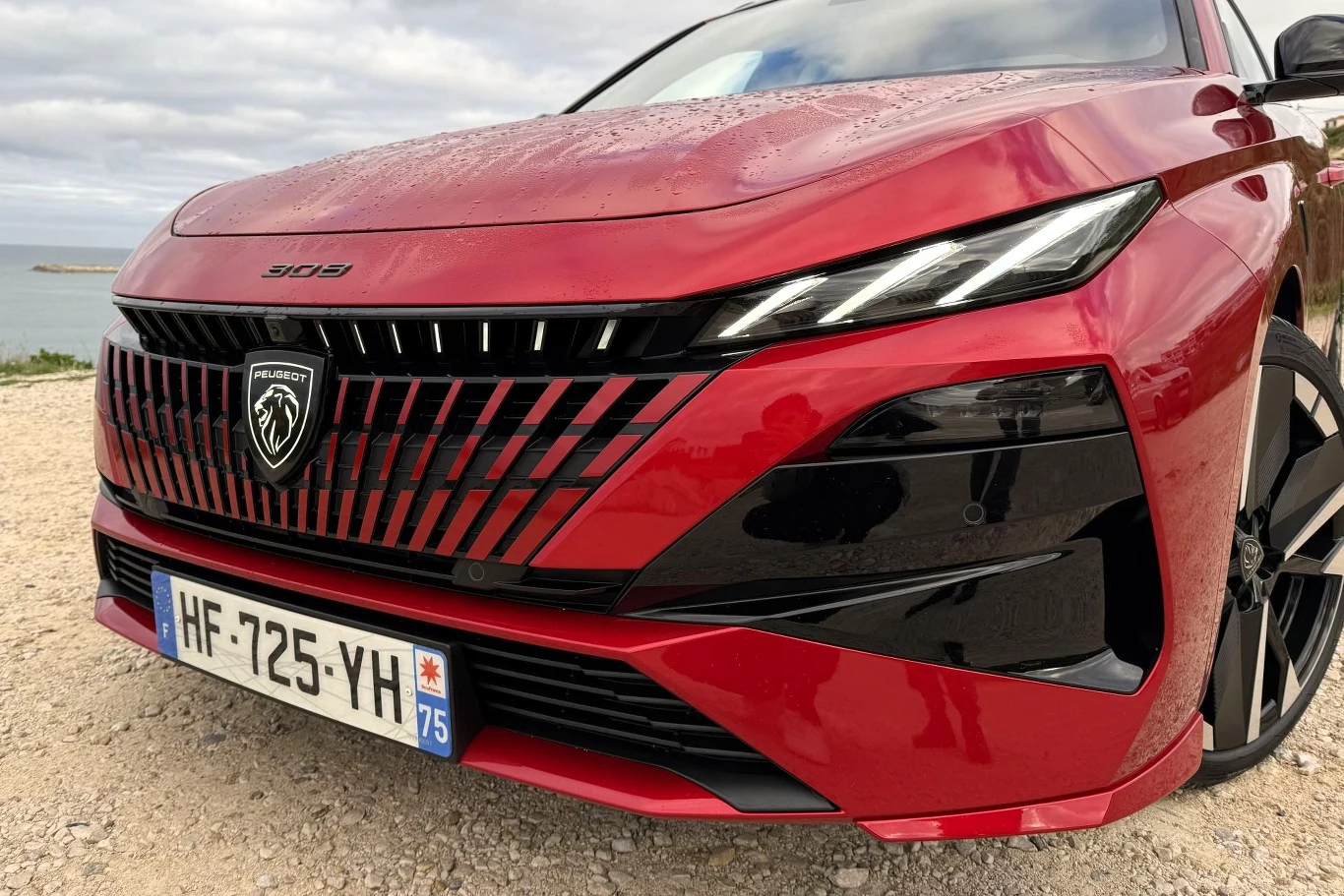 Czerwony samochód osobowy Peugeot 308 z wyraźnym, charakterystycznym grillem i nowoczesnymi reflektorami, widoczna francuska tablica rejestracyjna, auto stoi na żwirowym podłożu, w tle niebo z chmurami i fragment wody.