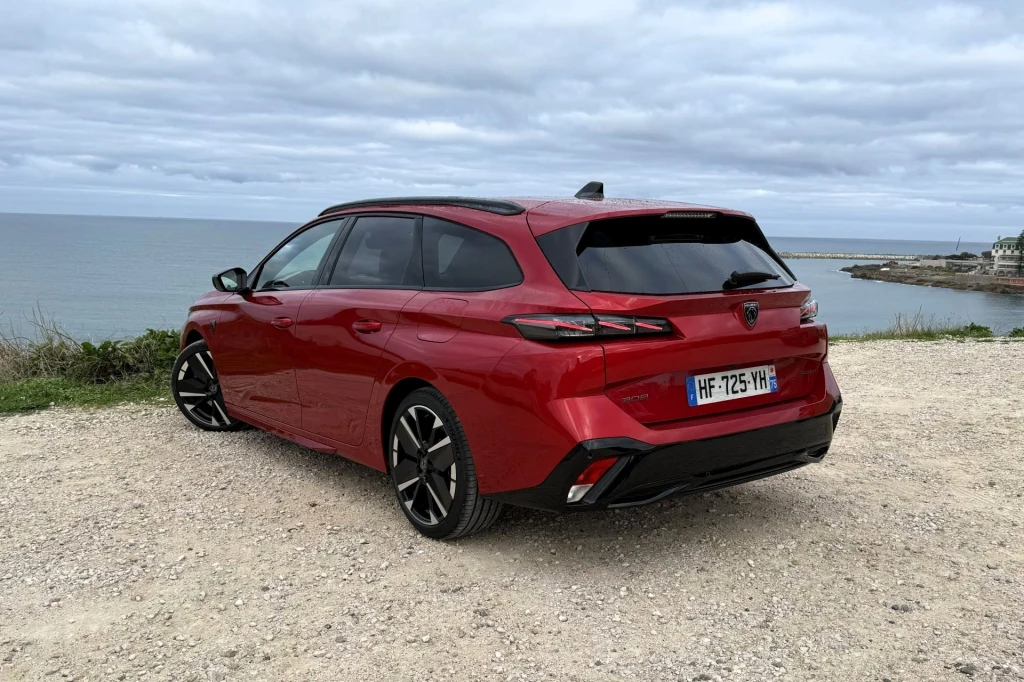 Czerwony samochód typu kombi o nowoczesnym designie zaparkowany na żwirowym terenie tuż przy wybrzeżu z widokiem na morze i efektownie ukształtowane chmury na niebie.