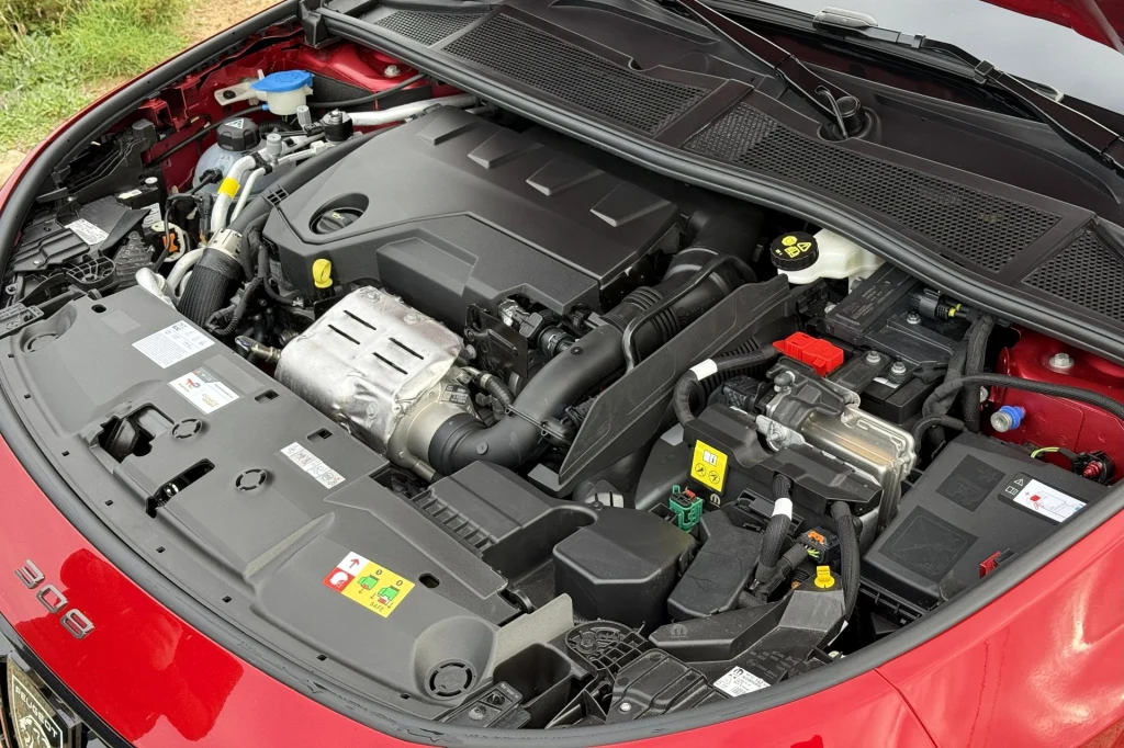 Komora silnika samochodu osobowego w kolorze czerwonym, widoczne liczne elementy mechaniczne, pokrywa silnika i akumulator, liczne przewody i oznaczenia bezpieczeństwa.