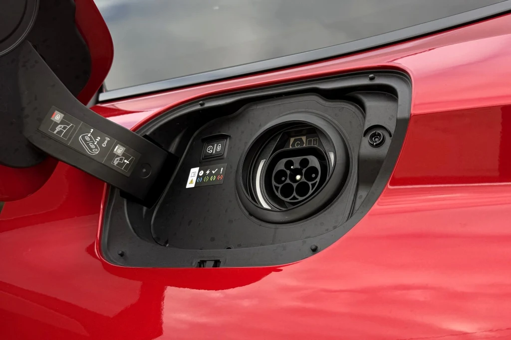 Gniazdo ładowania samochodu elektrycznego umieszczone na karoserii czerwonego auta, otwarta klapka ochronna i oznaczenia informacyjne ułatwiające prawidłowe podłączenie ładowarki.