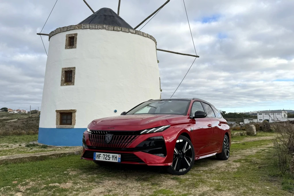 Czerwony samochód osobowy zaparkowany na tle tradycyjnego, białego wiatraka z niebieskim pasem i kamiennym dachem. Obok wiatraka widoczna jest otwarta przestrzeń oraz częściowo zachmurzone niebo.