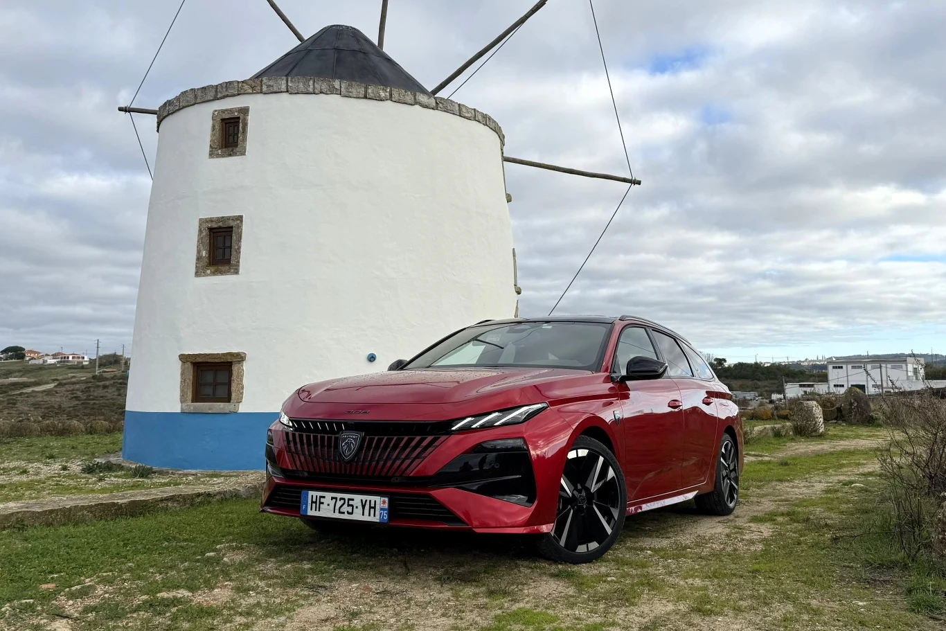 Czerwony samochód osobowy zaparkowany na tle tradycyjnego, białego wiatraka z niebieskim pasem i kamiennym dachem. Obok wiatraka widoczna jest otwarta przestrzeń oraz częściowo zachmurzone niebo.