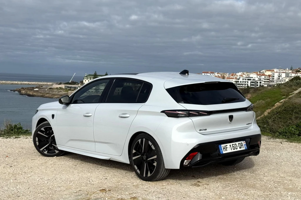 Biały samochód osobowy marki Peugeot stoi na żwirowej drodze z widokiem na wybrzeże i zabudowania miasteczka w tle, pod zachmurzonym niebem.