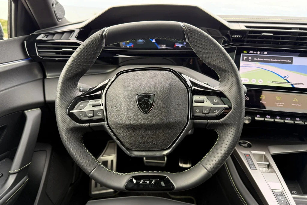 Nowoczesna kierownica samochodowa z logotypem producenta pośrodku, liczne przyciski do sterowania multimediów oraz wyświetlacz na desce rozdzielczej ze wskaźnikami i ekranem nawigacji.