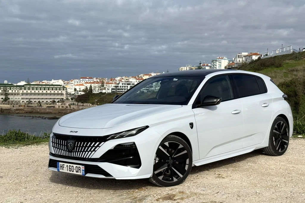 Nowoczesny, biały hatchback Peugeot stoi zaparkowany na tle malowniczej nadmorskiej panoramy z zabudowaniami i wybrzeżem pod zachmurzonym niebem.