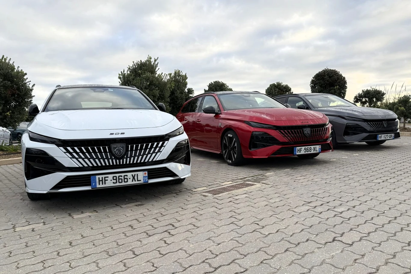 Trzy nowoczesne samochody marki Peugeot zaparkowane w rzędzie na parkingu, na pierwszym planie biały, obok czerwony oraz ciemnoszary, w tle niskie drzewa i pochmurne niebo.