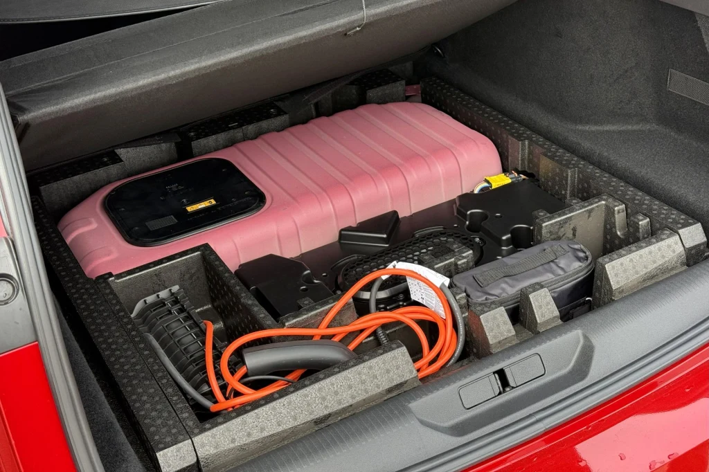 Bagażnik samochodu z widocznymi akcesoriami do ładowania, w tym różowawą walizką, przewodami elektrycznymi i elementami organizera z pianki zabezpieczającej. Wszystkie przedmioty uporządkowane w dedykowanych przegródkach.