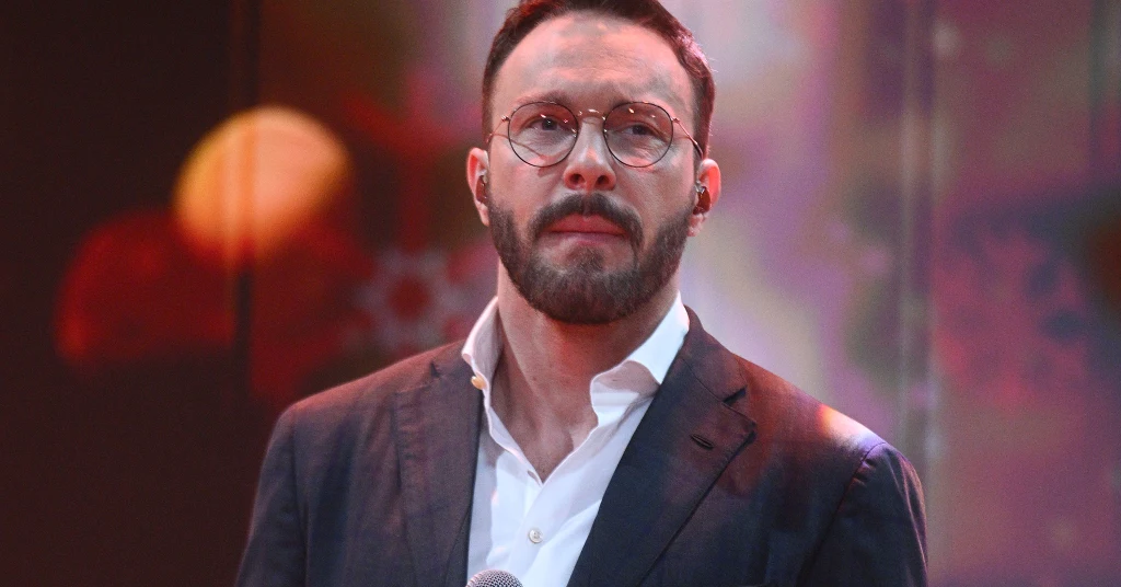 Sławek Uniatowski Mężczyzna w garniturze i białej koszuli stoi na scenie podczas występu, trzymając mikrofon, w tle widoczne kolorowe rozmyte światła.