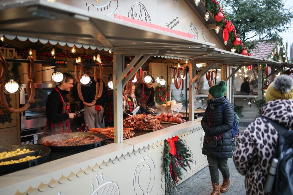 Stoisko na jarmarku świątecznym z różnymi potrawami oraz wyrobami mięsnymi, obsługiwane przez osoby w fartuchach w kratkę. Klientka w ciepłej kurtce i zielonej czapce zamawia jedzenie, wokół dekoracje świąteczne.
