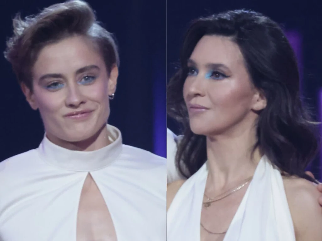 Dwie kobiety w białych strojach scenicznych ze starannie ułożonymi włosami i wyrazistym makijażem, stojące obok siebie na tle ciemnego, podświetlonego światłem reflektorów tła.